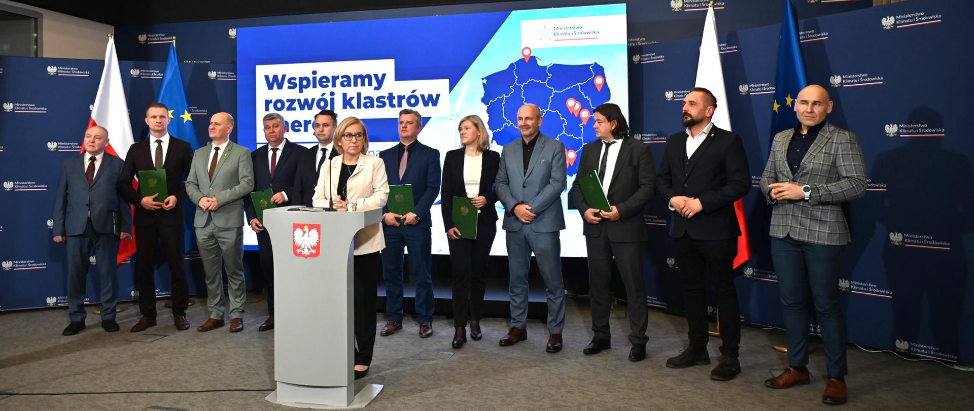 Na środku, za mównicą stoi ministra klimatu i środowiska Paulina Hennig-Kloska. Za nią stoi 11 przedstawicieli klastrów energii, czyli przedstawicieli lokalnych inicjatyw zrzeszających samorządy, przedsiębiorstwa i instytucje działające na rzecz rozwoju energetyki rozproszonej i odnawialnych źródeł energii.