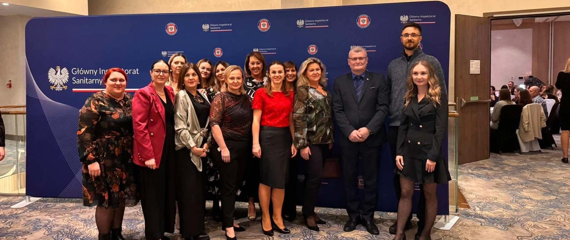 Grupa kilkunastu uczestników Forum Wiedzy Państwowej Inspekcji Sanitarnej stoi na tle granatowej ścianki z logotypami Głównego Inspektoratu Sanitarnego. Osoby ustawione są w dwóch rzędach, uśmiechają się do zdjęcia w eleganckich, konferencyjnych strojach. W tle widoczne jest wejście do sali konferencyjnej.