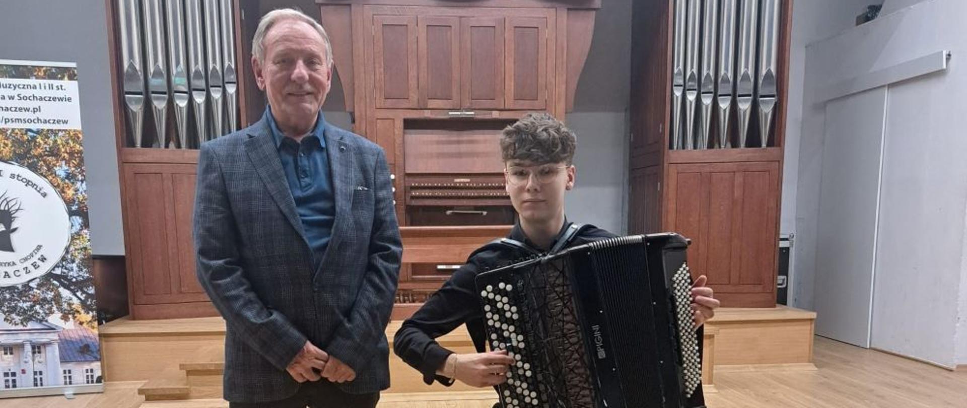 Zdjęcie kolorowe – Od lewej stoi nauczyciel akordeonu Zbigniew Stawiarz w ciemnym garniturze, a obok na krześle siedzi Stanisław Rogala z akordeonem na kolanach w czarnej koszuli i czarnych spodniach.