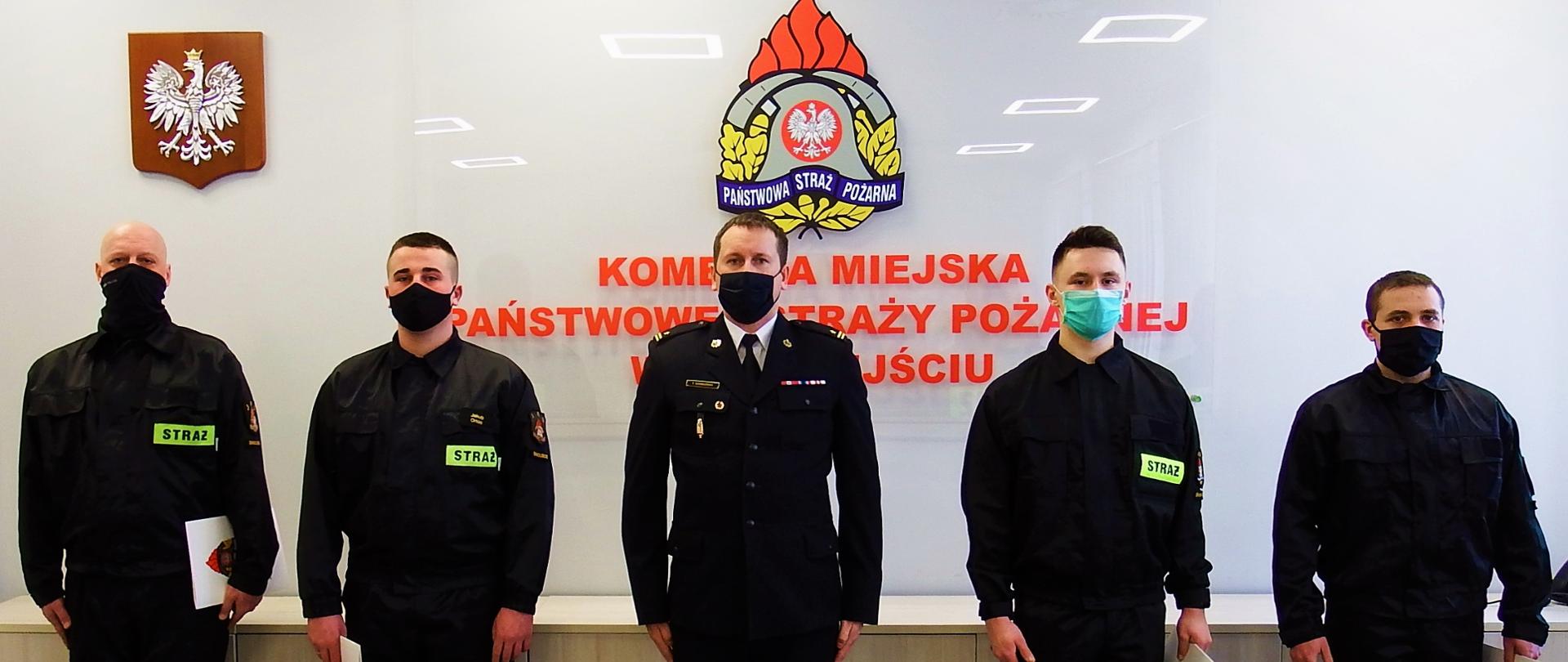 Pięciu strażaków en face na tle godła państwowego i godła PSP. Od lewej str. Mariusz Junik, str. Jakub Orlon, mł. bryg. Tomasz Samborski, str. Tobiasz Flaczyński, str. Krzysztof Fabiańczuk. Strażacy w mundurach koszarowych, oficer w mundurze wyjściowym.