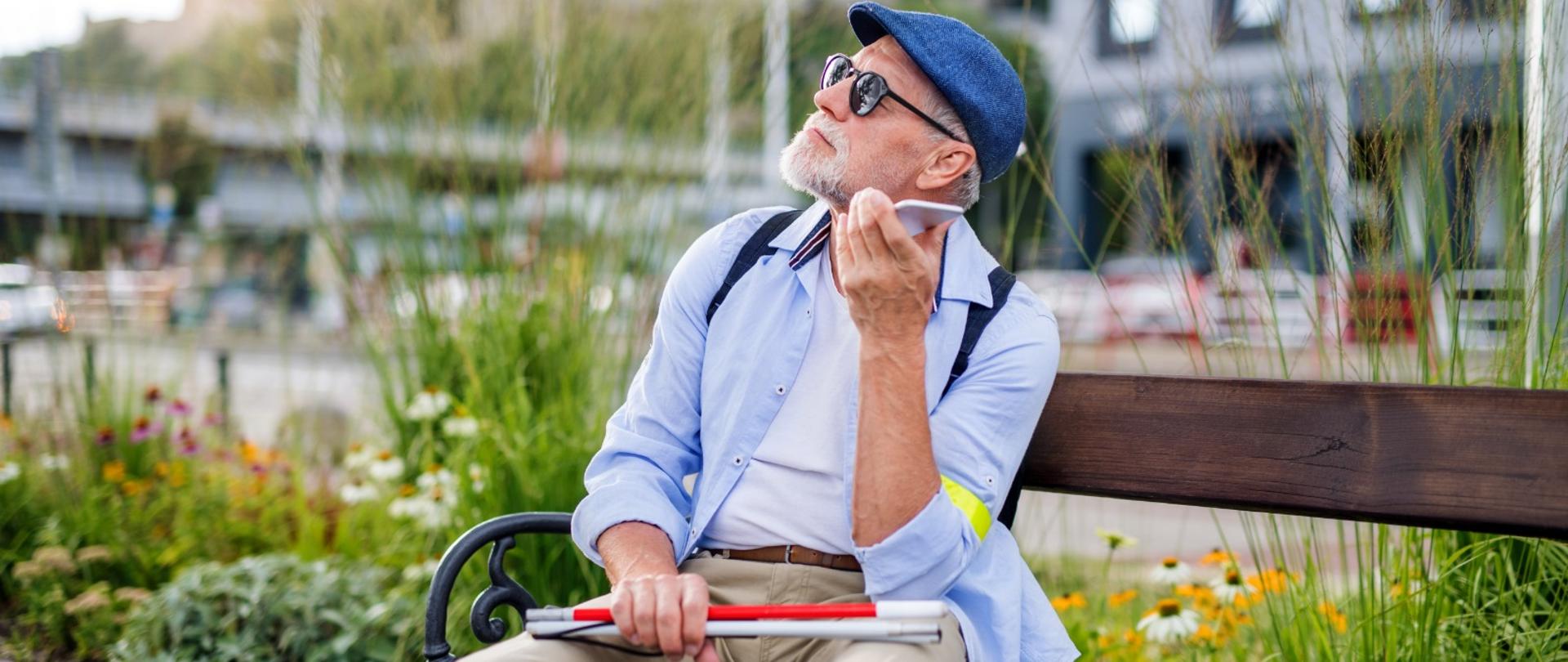 Na zdjęciu niewidomy mężczyzna trzymający przy uchu smartfon. Mężczyzna siedzi na ławce, w tle widać kwiaty i rośliny. Na dalszym planie rozmyte budynki. Ubrany jest w jasne spodnie, błękitną koszulę i niebieski kaszkiet. W dłoni trzyma złożoną białą laskę.