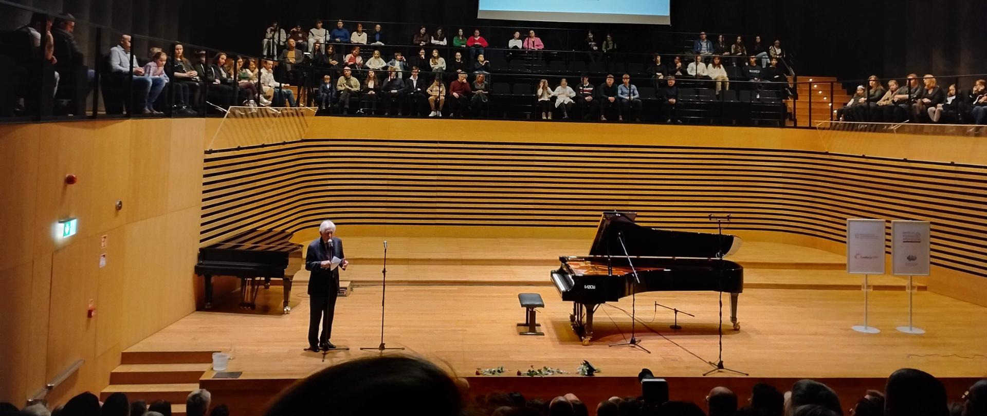 Zdjęcie przedstawia salę koncertową PSM w Jastrzębiu Zdroju, na środku sceny stoi fortepian, po lewej prelegent przy mikrofonie, nad sceną siedzi publiczność