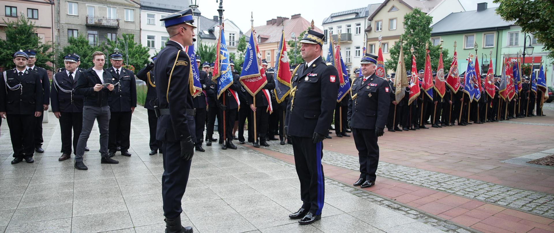 Na miejskim placu strażak w galowym mundurze stoi na baczność naprzeciw pocztów sztandarowych ustawionych w szeregu. W tle zgromadzili się liczni funkcjonariusze oraz mieszkańcy uczestniczący w obchodach. Plac otaczają drzewa i zabudowa miejska, a pogoda jest pochmurna, lecz bez intensywnych opadów.
