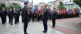 Na miejskim placu strażak w galowym mundurze stoi na baczność naprzeciw pocztów sztandarowych ustawionych w szeregu. W tle zgromadzili się liczni funkcjonariusze oraz mieszkańcy uczestniczący w obchodach. Plac otaczają drzewa i zabudowa miejska, a pogoda jest pochmurna, lecz bez intensywnych opadów.