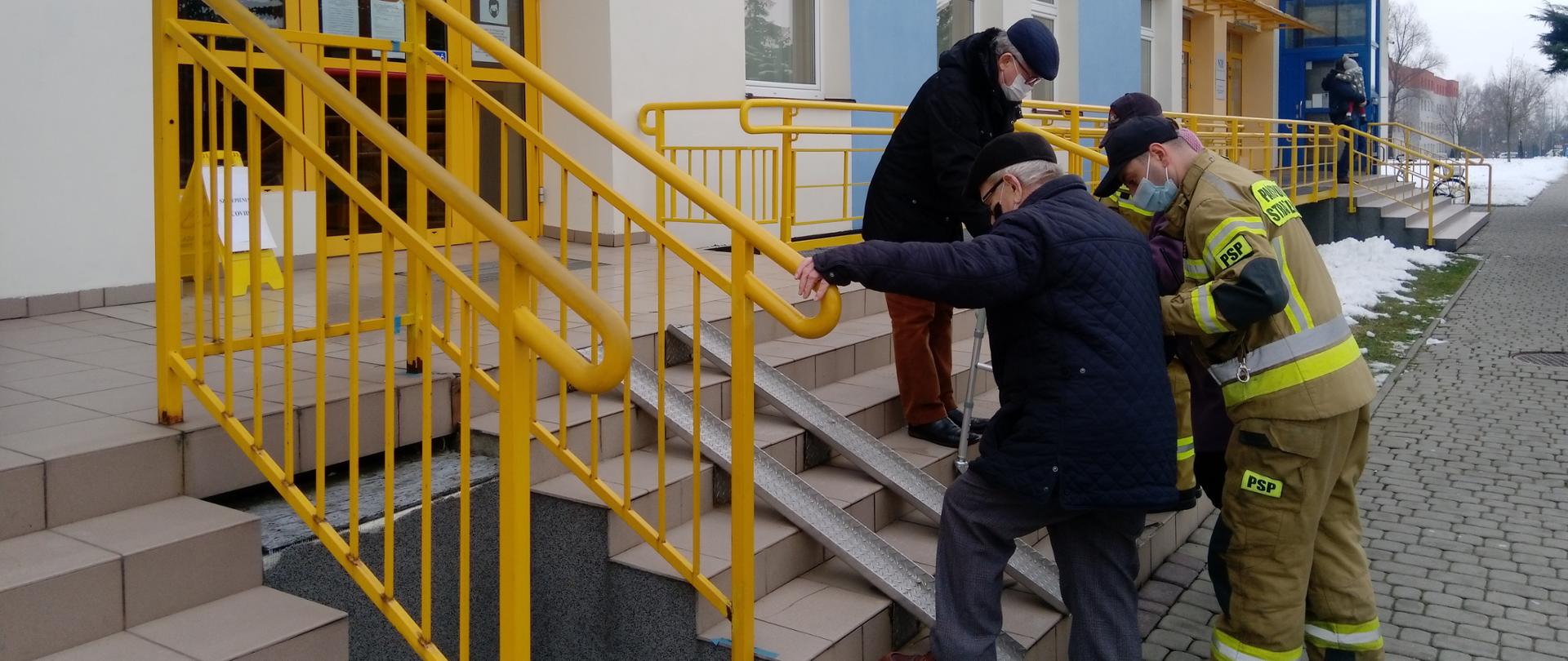 Osoby w podeszłym wieku przy pomocy strażaków wchodzą po schodach do przychodni lekarskiej. W tle kolorowa elewacja budynku.