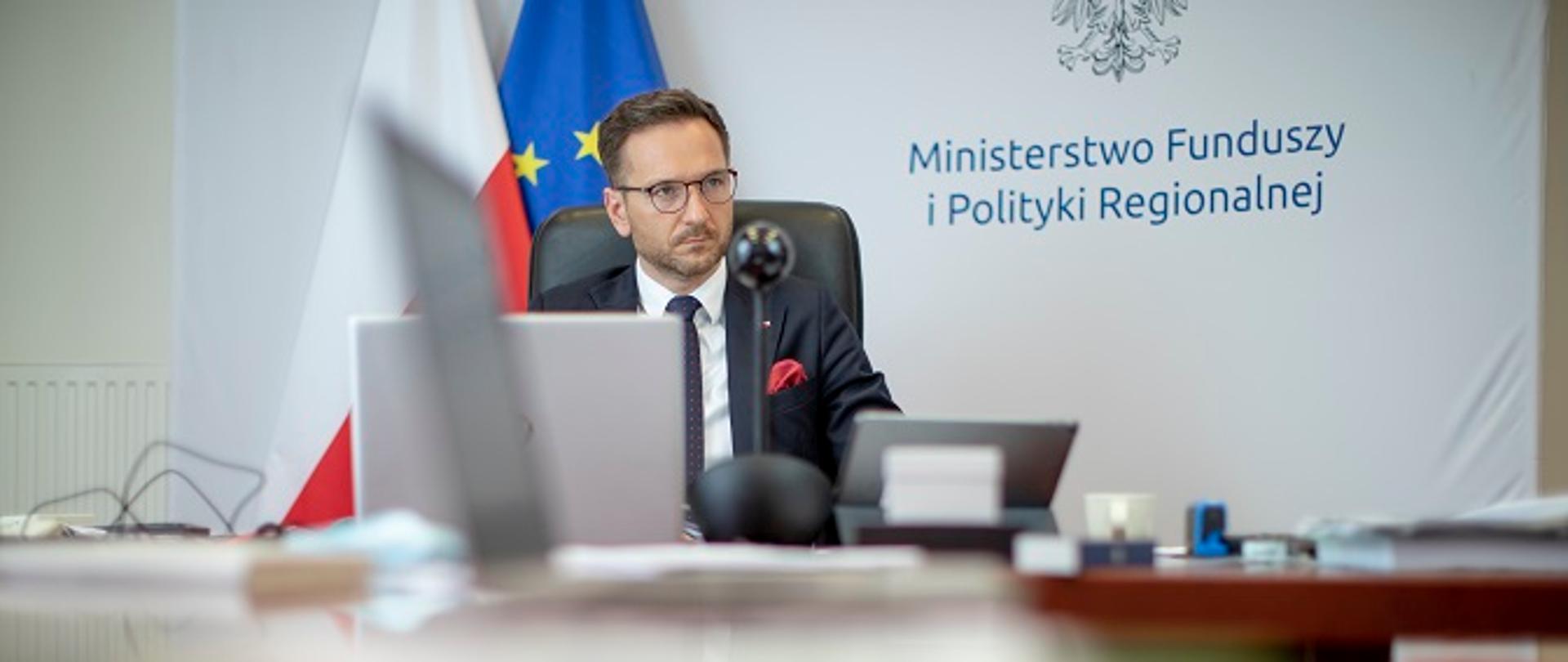 Minister Waldemar Buda siedzi za stołem konferencyjnym, przed nim laptop, w tle ścianka z log MFiPR