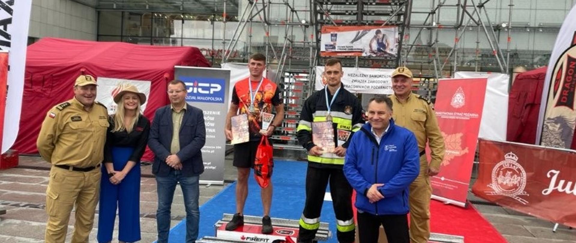 Na zdjęciu widoczne w centralnej części fotografii dwie osoby z medalami oraz dyplomami. Przed nimi prądownica, węże oraz młoty wykorzystywane w zawodach. Po prawej stoją dwie osoby, a po lewej trzy. Na drugim planie widoczna konstrukcja na której odbywały się zawody z napisem „Komenda Miejsca PSP w Krakowie”.