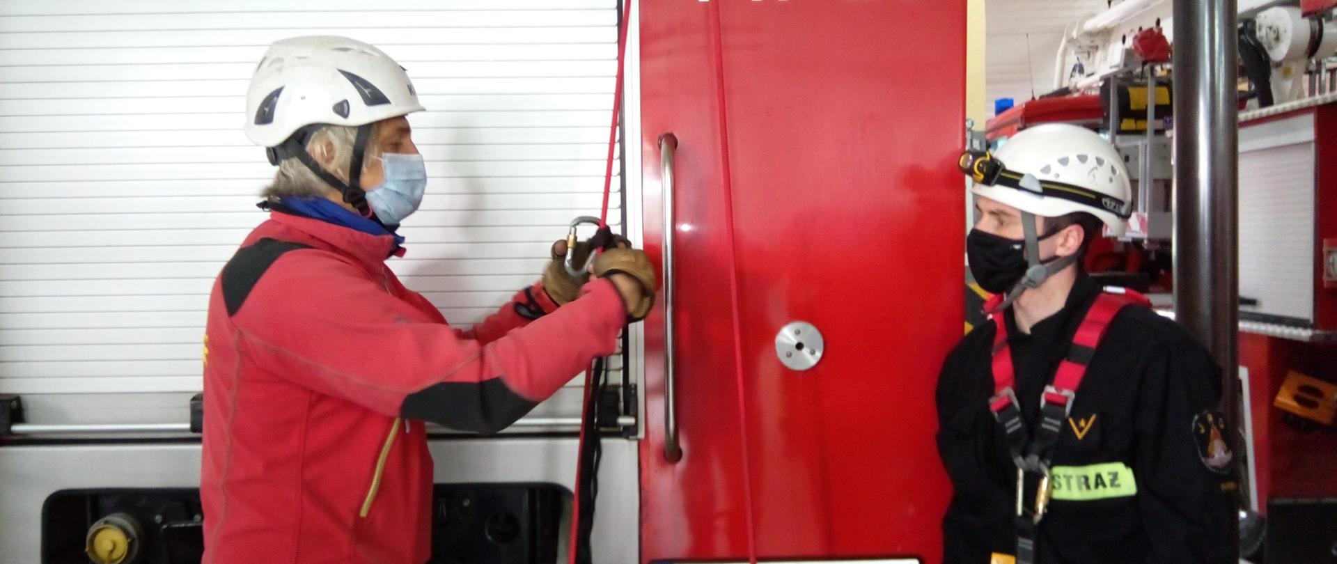 Instruktor ratownictwa wysokościowego ubrany w czerwoną bluzę polarową i biały kask objaśnia umundurowanemu ratownikowi techniki wiązania węzłów ratowniczych. 