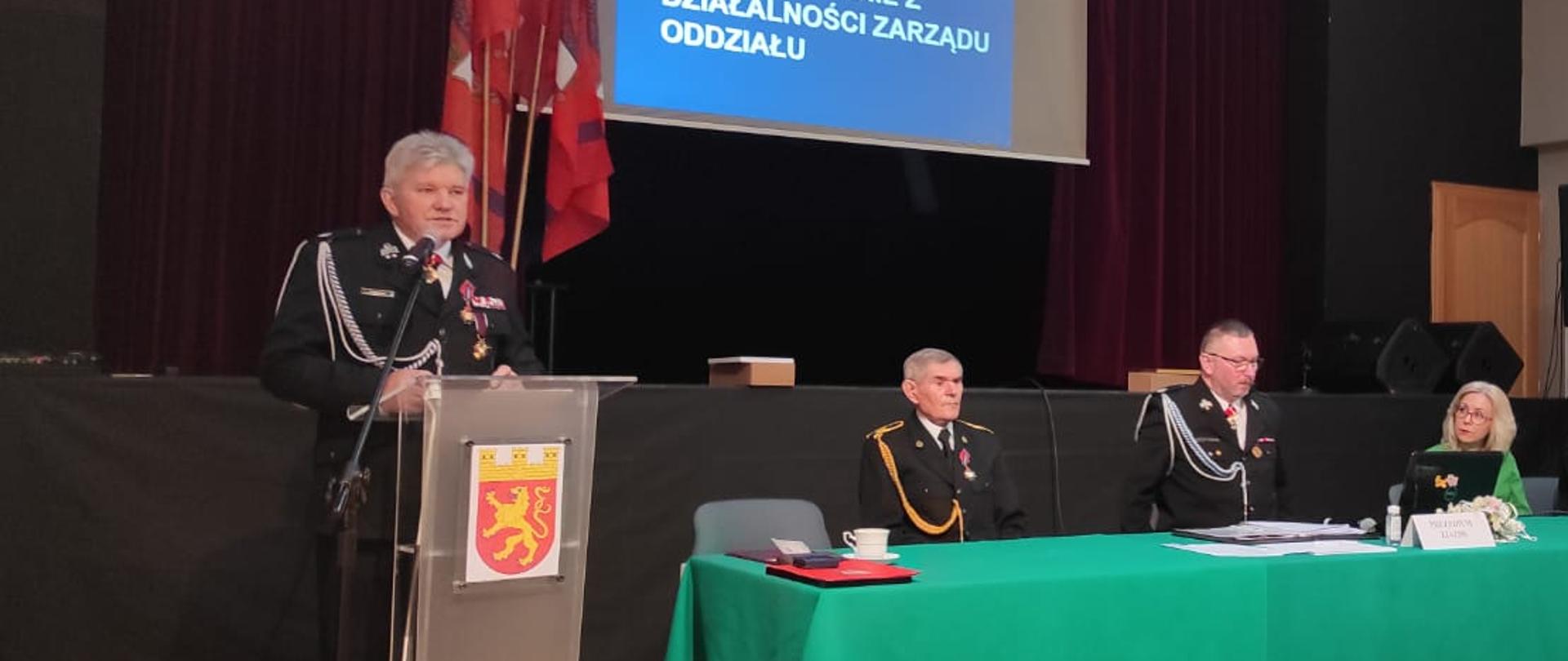 Strażak stoi przy mównicy, obok kilkoro strażaków siedzi przy długim stole prezydialnym, na którym rozłożony jest zielony obrus. W tle projektor wyswietla prezentacje.