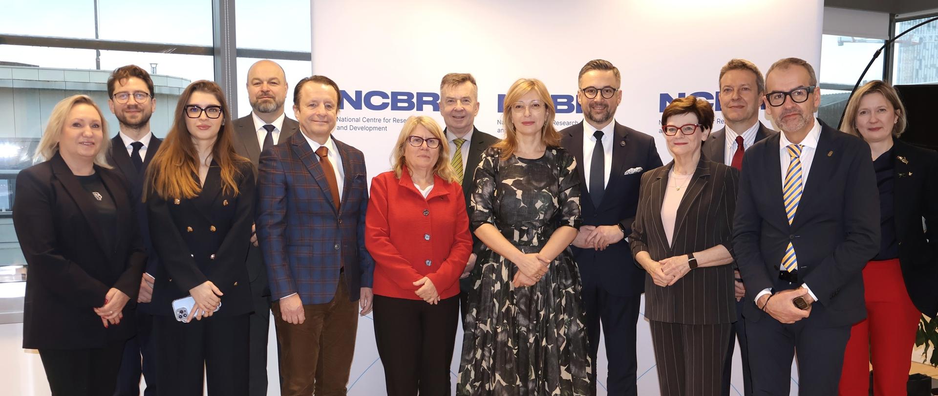 Zdjęcie zbiorowe, pod ścianką z napisem NCBR stoi kilkanaście osób, pośrodku grupy minister Kulasek i kobieta w sukience w biało-czarny wzorek.