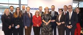 Zdjęcie zbiorowe, pod ścianką z napisem NCBR stoi kilkanaście osób, pośrodku grupy minister Kulasek i kobieta w sukience w biało-czarny wzorek.