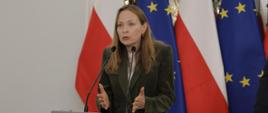 Minister funduszy i polityki regionalnej Katarzyna Pełczyńska-Nałęcz stoi za mównicą z mikrofonami, za jej plecami flagi Polski i UE