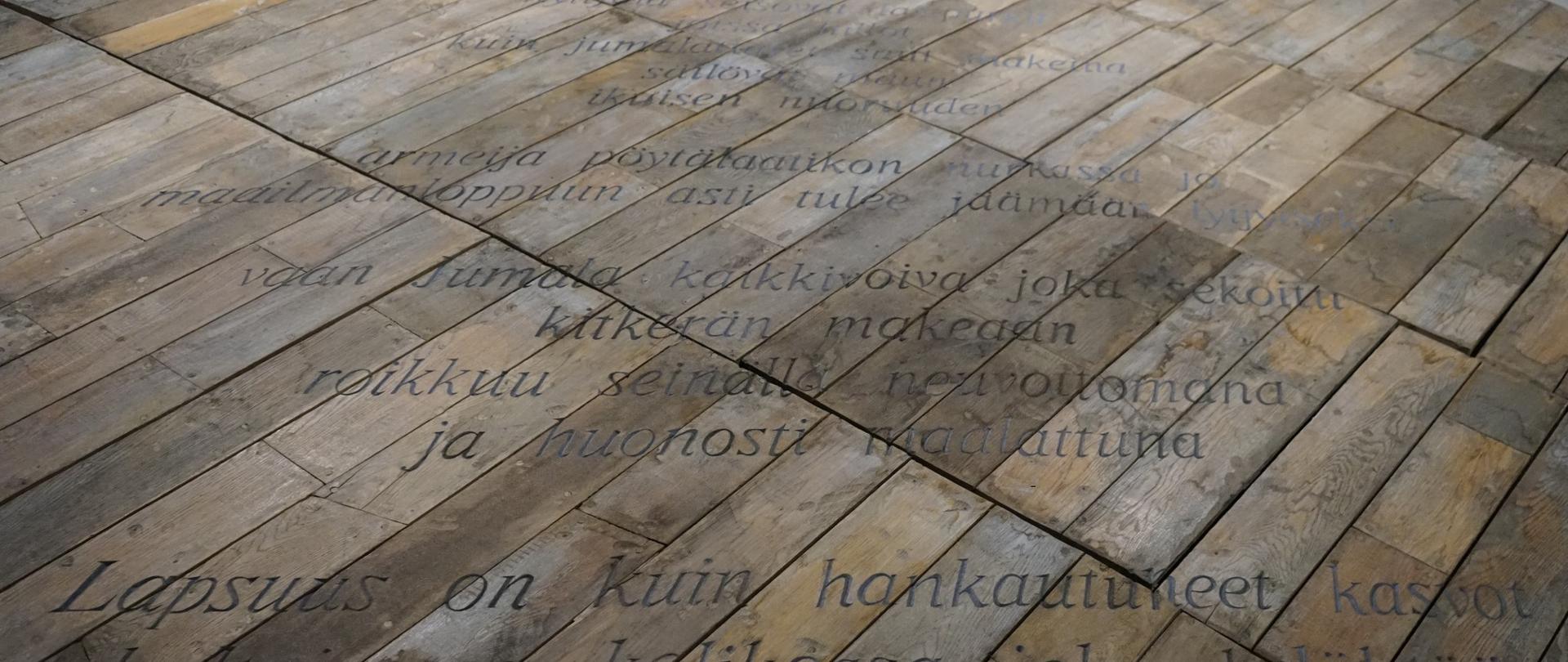Deski ułożone są nierówno, odstają. Przypominają stary parkiet. Deski są zniszczone, odbarwione, w różnych kolorach brązu i szarości. Na nich znajdują się napisy, prawdopodobnie w języku fińskim.
