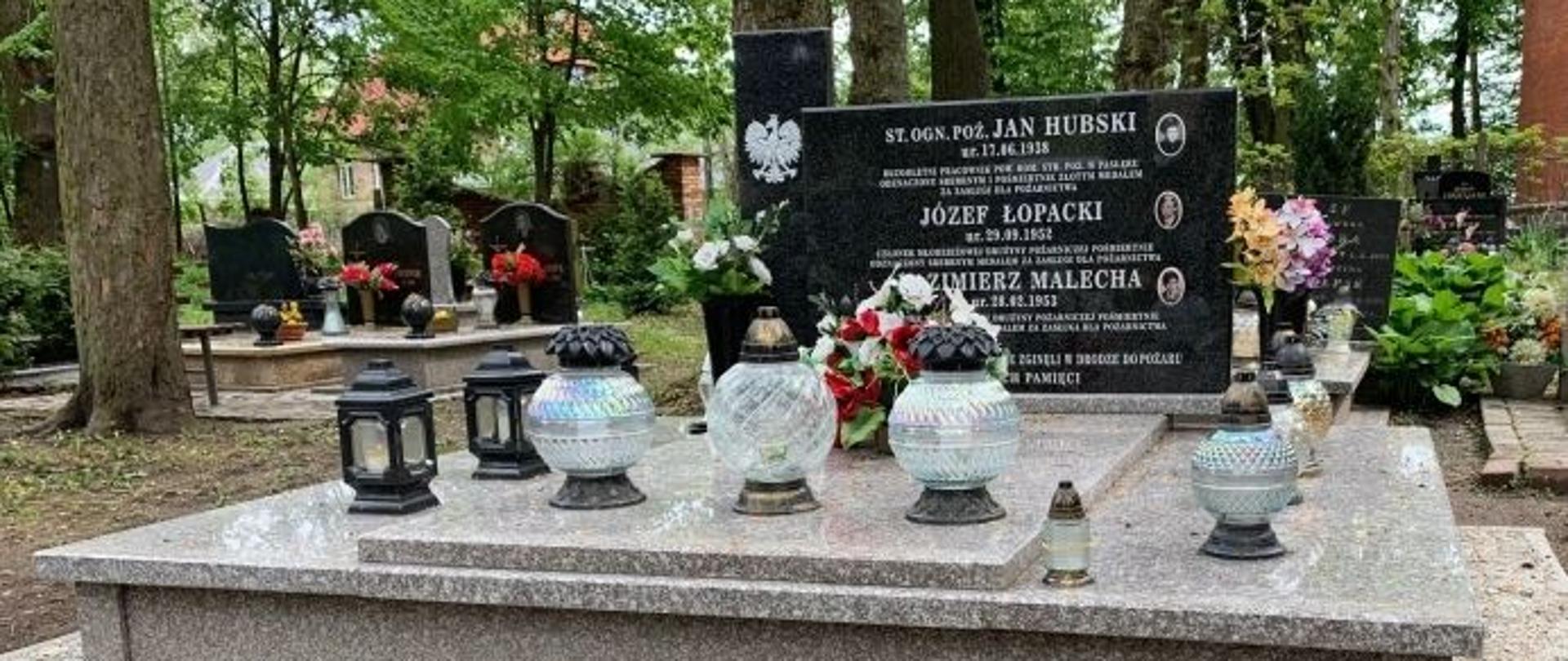 Zdjęcie przedstawia zbiorowy pomnik na cmentarzu, w którym pochowano zmarłych tragicznie strażaków. W tle widać drzewa oraz inne groby zmarłych.