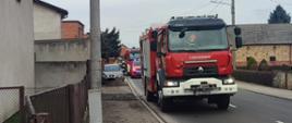 Samochody strażackie stoją w szeregu na jezdni obok widoczne budynki mieszkalne.