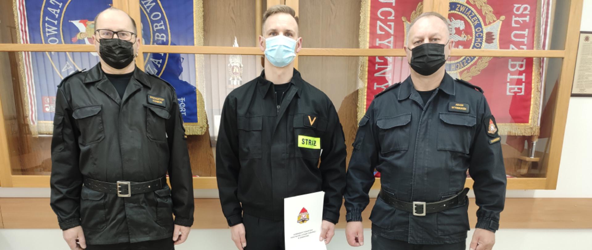 Zdjęcie przedstawia od lewej Komendanta Powiatowego Państwowej Straży Pożarnej w Zambrowie starszego brygadiera Krzysztofa Konieckiego w ubraniu dowódczo-sztabowym, ogniomistrza Damiana Dębka w ubraniu koszarowym oraz Zastępcę Komendanta Powiatowego Państwowej Straży Pożarnej w Zambrowie młodszego brygadiera Adama Wysmułka w ubraniu dowódczo-sztabowym. Zdjęcie zostało zrobione z okazji mianowania na wyższe stanowisko służbowe ogniomistrza Damiana Dębka i wykonane w jednym z pomieszczeń Komendy Powiatowej Państwowej Straży Pożarnej w Zambrowie.