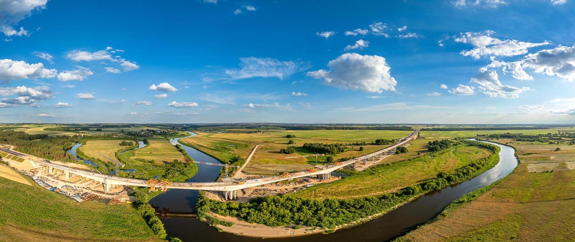Widok z lotu ptaka na wielką plac budowy mostu nad meandrującą rzeką. Rzeka wygina się przez krajobraz, zielone pola i skupiska drzew po obu stronach. Na placu budowy widać wiele żurawi, kilka częściowo ukończonych odcinków mostu z widocznymi rusztowaniami oraz liczne pojazdy budowlane. Niebo jest bezchmurne, co sugeruje dobre warunki pogodowe do prac budowlanych.
