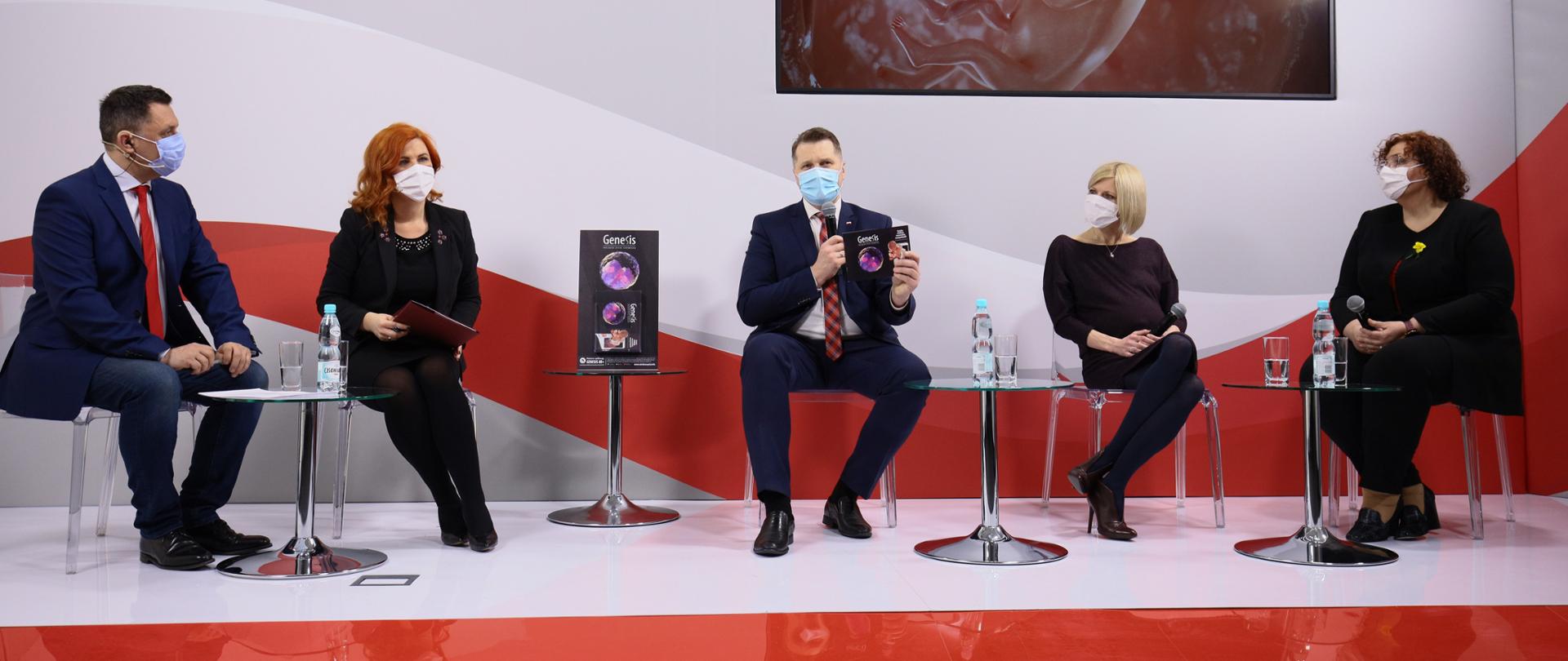 Pięć osób siedzi na krzesłach na biało-czerwonej scenie. Dwie osoby to mężczyźni ubrani w garnitury, pozostałe 3 to ubrane na ciemno kobiety. Jedna z kobiet jest w ciąży. Pomiędzy osobami stoją szklane stoliki. Na stolikach szklanki oraz wody. Pośrodku minister Przemysław Czarnek z mikrofonem oraz książeczką Genesis w ręku. Tłem sceny jest logo Ministerstwa Edukacji i Nauki oraz falująca biało-czerwona flaga. W tle także monitor z napisem Genesis. Początek życia człowieka Narodowy Dzień Życia 24 marca.
