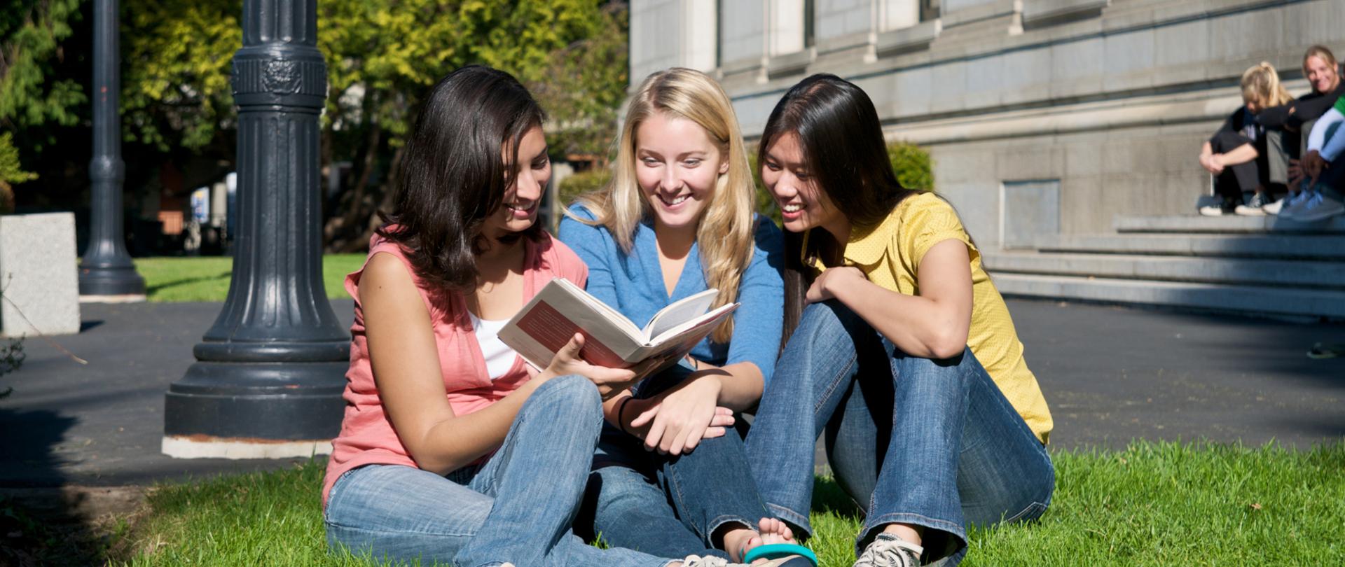 Trzy młode osoby siedzą na trawniku pod budynkiem uczelni.