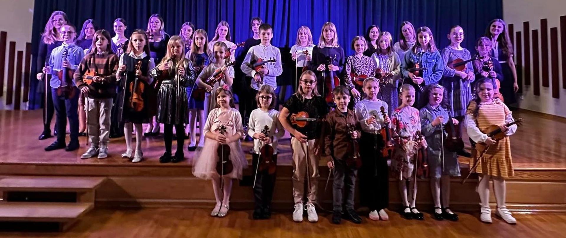 Uczestnicy spotkania stoją trzymając instrumenty w 3 rzędach na scenia.