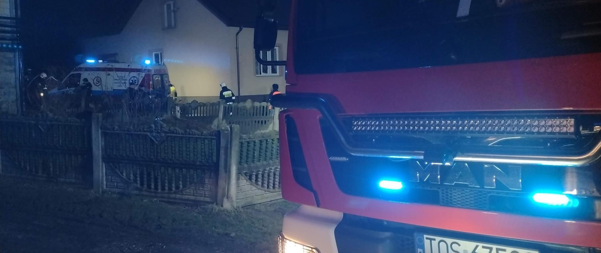 Na pierwszym planie samochód gaśniczy. W tle budynek w którym doszło do pożaru, a obok karetka ratownicy i strażacy.