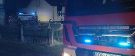 Na pierwszym planie samochód gaśniczy. W tle budynek w którym doszło do pożaru, a obok karetka ratownicy i strażacy.