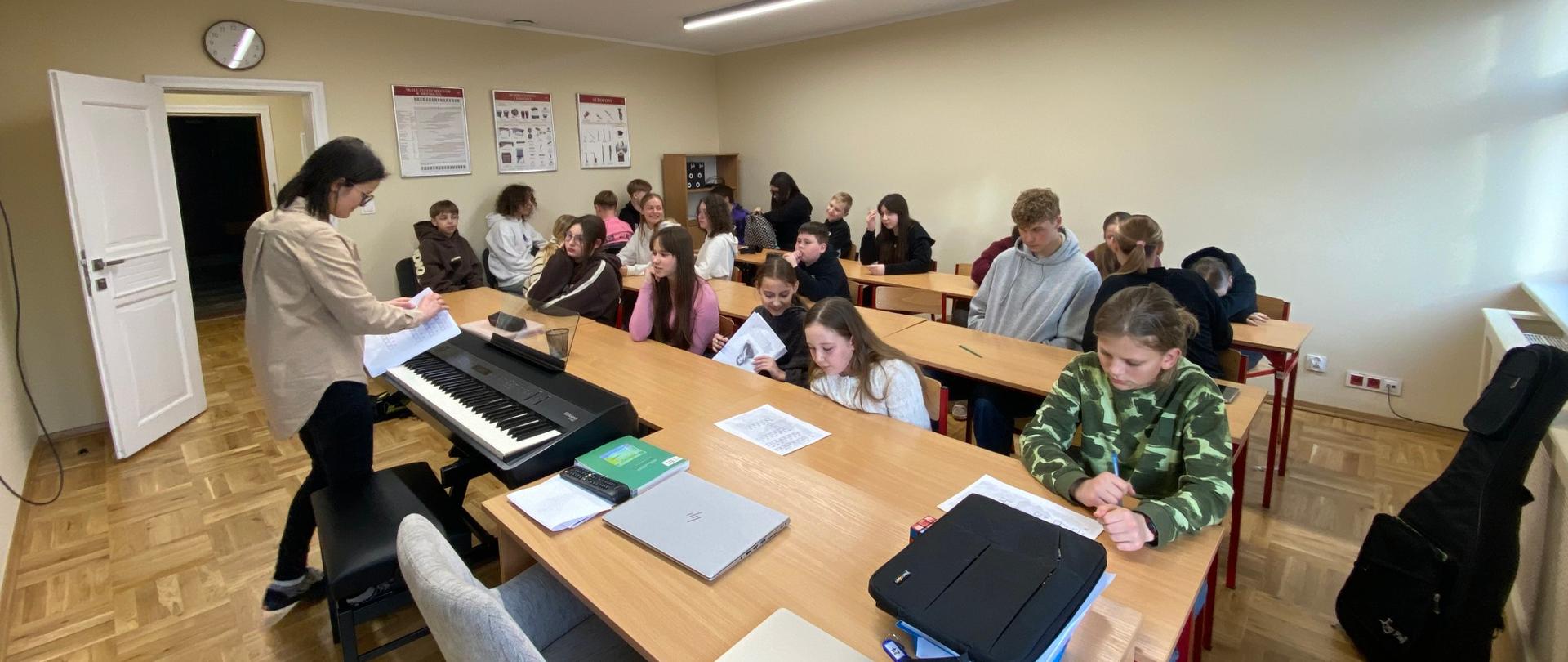 Uczniowie siedzący w ławkach w sali lekcyjnej, nauczycielka stojąca przy pianinie elektronicznym.