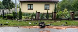 Uszkodzone ogrodzenie posesji oraz szczątki rozbitego samochodu znajdujące się na drodze