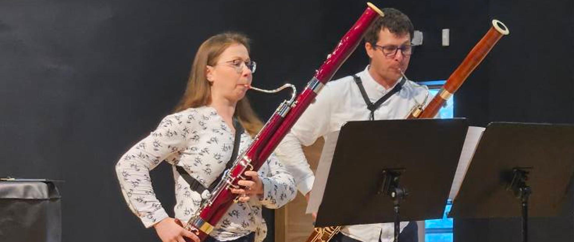 Kolorowe zdjęcie, na który znajdują się dwie osoby grające na fagotach. Osoby znajdują się na scenie przed nimi stoją dwa czarne pulpity z nutami. 