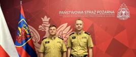 Dwóch mężczyzn w beżowych mundurach Państwowej Straży Pożarnej stoi obok siebie na tym samym tle z czerwonym napisem i symbolicznym orłem. Po lewej stronie znajdują się flaga Polski i flaga z gryfem. Jeden z mężczyzn trzyma w lewej ręce biały dokument.