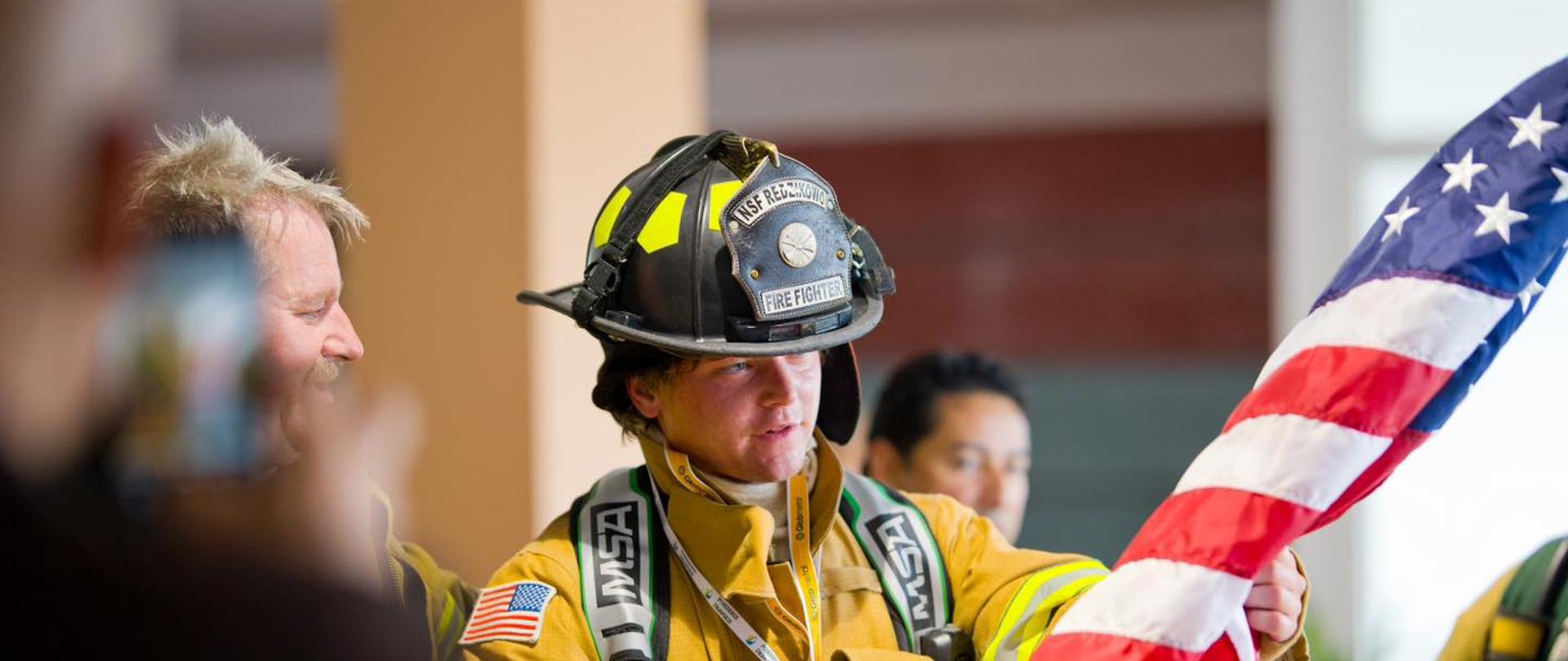 Na zdjęciu znajduje się strażak w pełnym rynsztunku. Ma na sobie grubą, żółtą kurtkę, czarne spodnie i kask, na którym znajduje się plakietka z napisem „FIRE FIGHTER”. Ubranie strażaka jest częściowo osłonięte przez flagę Stanów Zjednoczonych, którą trzyma w rękach. Strażak ma lekko rozczochrane, brązowe włosy i uśmiecha się. W tle widać innych strażaków i flagę Stanów Zjednoczonych.