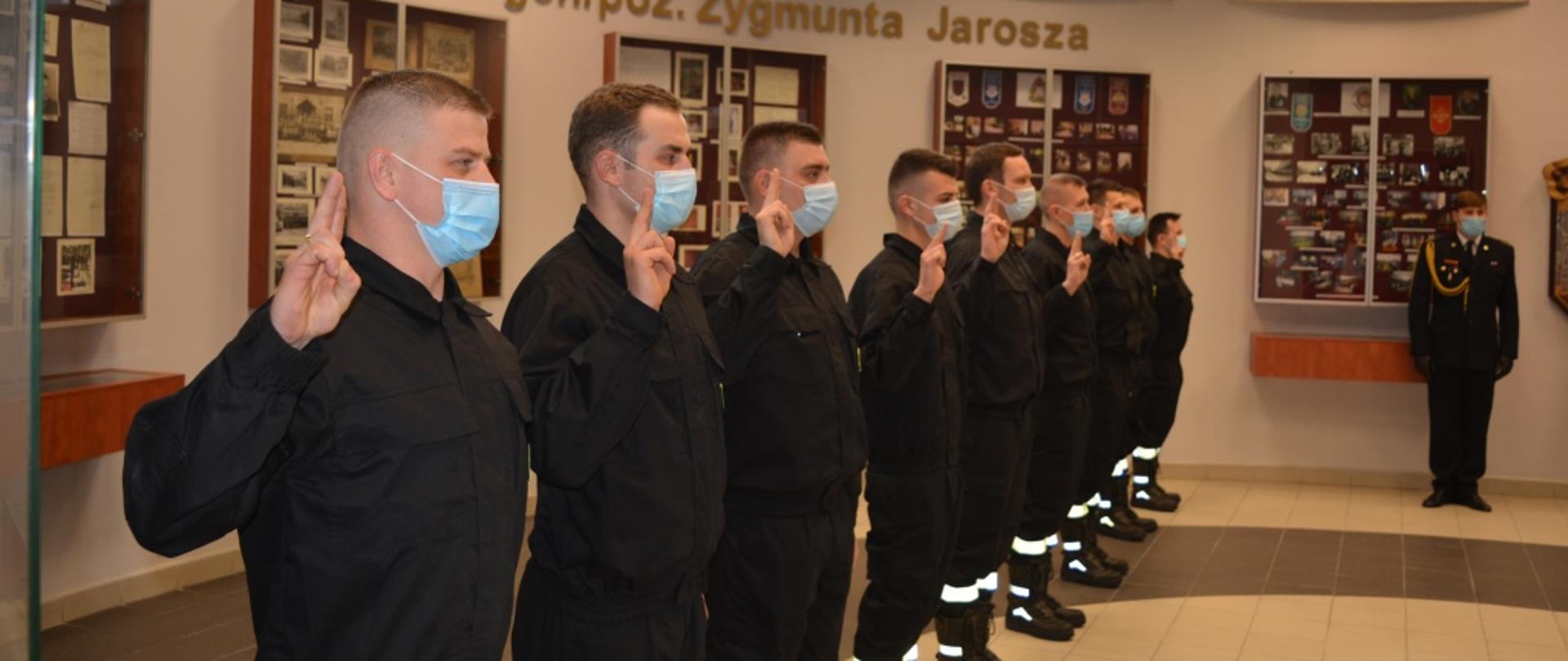 Zdjęcie przedstawia moment ślubowania nowo przyjętych funkcjonariuszy. Strażacy ustawieni są w rzędzie jeden obok drugiego. Ubrani są w czarne ubrania koszarowe z jaskrawymi elementami odblaskowymi. Prawą rękę trzymają podniesioną w górę na znak ślubowania. 