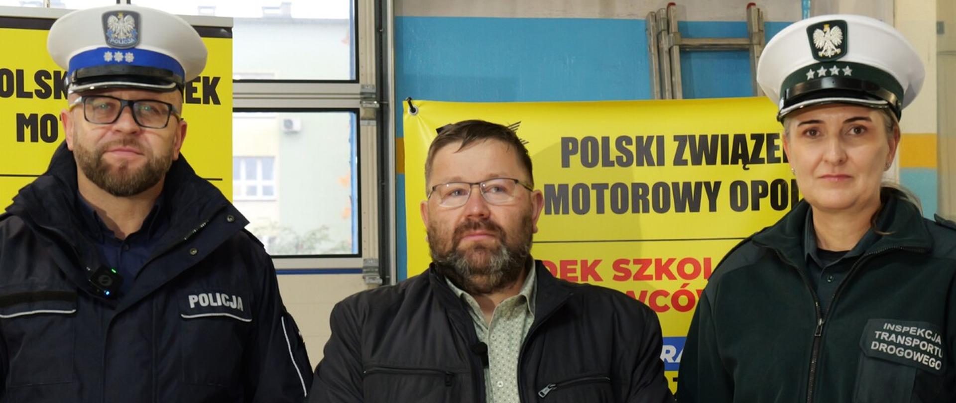 Trzy osoby stojące obok siebie na tle żółtych banerów Polskiego Związku Motorowego: policjant w mundurze i czapce z niebieskim otokiem, mężczyzna w ciemnej kurtce i okularach oraz funkcjonariuszka Inspekcji Transportu Drogowego w zielonym mundurze.