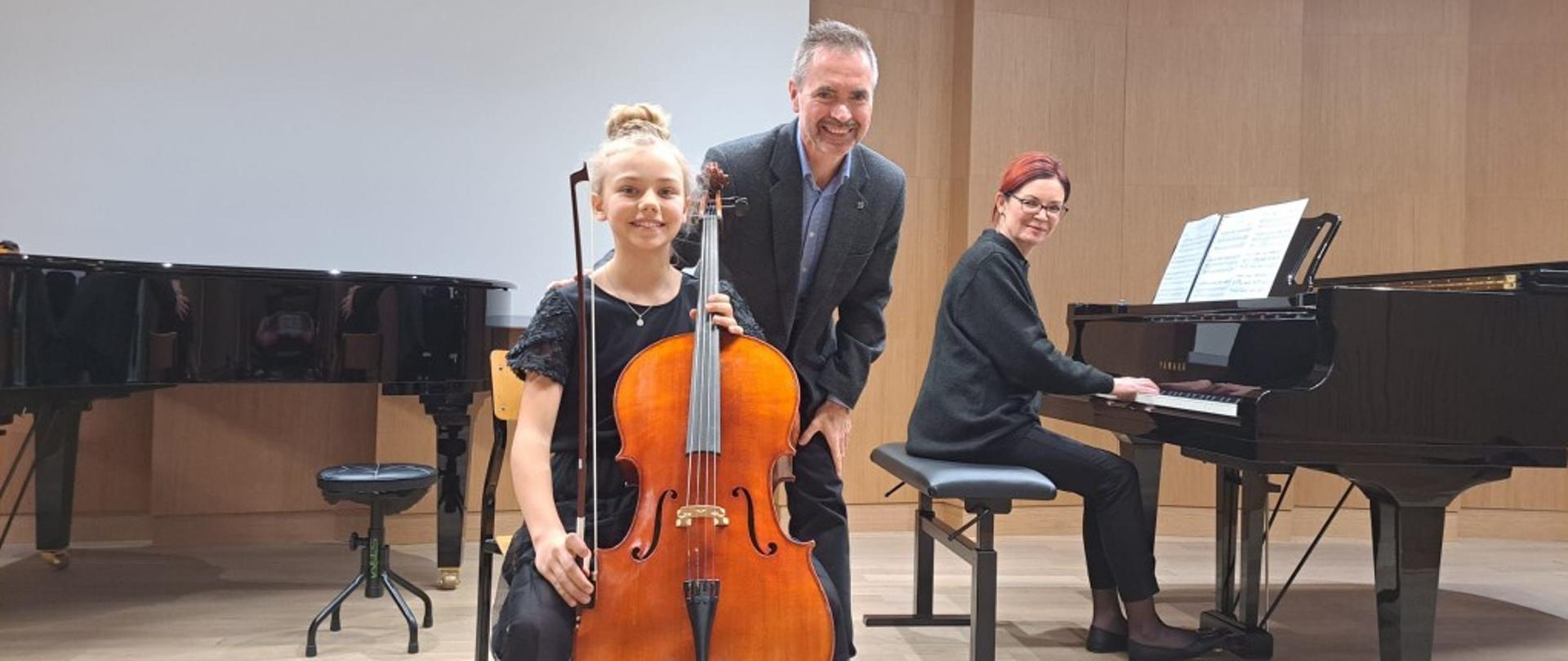 Na krześle siedzi dziewczynka w czarnej sukience z wiolonczelą. Za nią stoi nauczyciel. Przy fortepianie siedzi akompaniatorka.