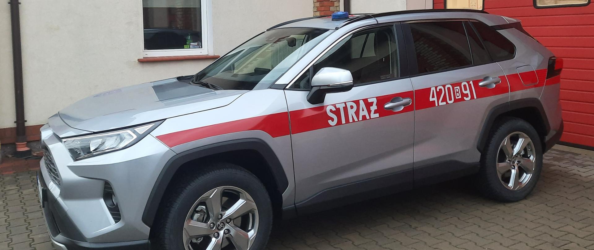 Na zdjęciu samochód operacyjny SLOp Toyota Rav 4 w kolorze srebrnym z czerwonym pasem i napisem straż na boku pojazdu. W tle budynek Komendy Powiatowej PSP w Grajewie.