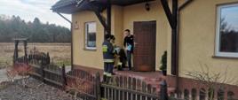 Zdjęcie przedstawia moment wręczenia ulotek przez dwóch strażaków mężczyźnie, który stoi w drzwiach domu jednorodzinnego.