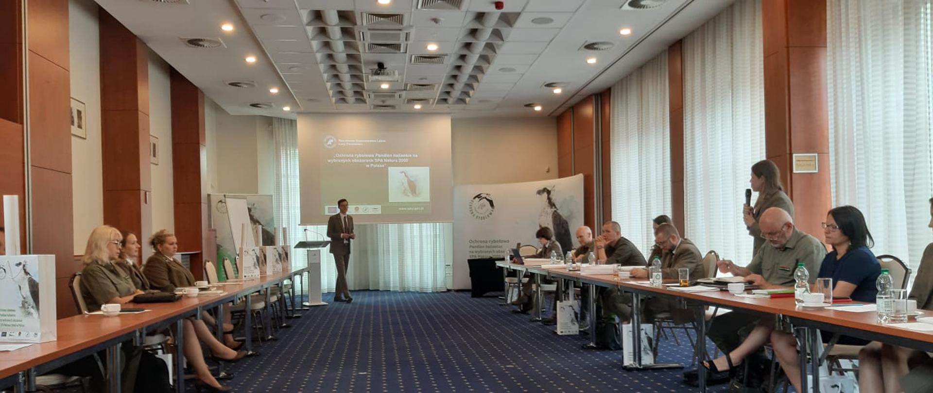 Sala wypełniona uczestnikami spotkania. Po lewej stronie trzy kobiety siedzą przy stole, po prawej stronie osiem osób przy stole. Jedna osoba stoi i mówi przez mikrofon. Na środku mężczyzna za nim wyświetla się temat spotkania. Obok po prawej stronie baner rybołowa. Na podłodze niebieski dywan