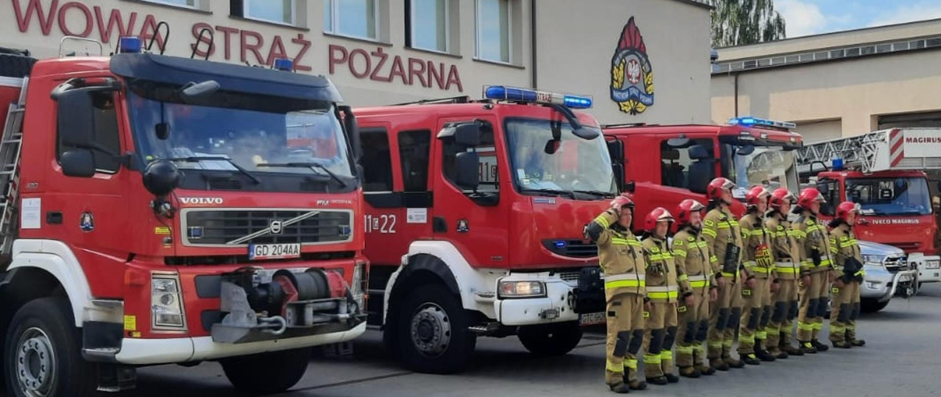 Ośmiu strażaków w ubraniach specjalnych stoi na baczność w mundurach specjalnych i hełmach. Pierwszy w szeregu dowództwa oddaje honor. Za nimi samochody ratowniczo-gaśnicze, za samochodami budynek jednostki.