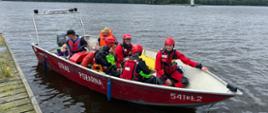 Na zdjęciu widoczna jest czerwona łódź motorowa należąca do straży pożarnej w Szczecinku („STRAŻ POŻARNA”), przycumowana do drewnianego pomostu nad jeziorem. Na pokładzie znajduje się kilku ratowników wodnych w czerwonych i zielonych kombinezonach ratowniczych oraz kaskach ochronnych. Jeden z ratowników siedzi na dziobie łodzi, a pozostali są rozmieszczeni w różnych częściach jednostki, część z nich uśmiecha się i rozmawia. W łodzi znajdują się również dwie osoby w pomarańczowych kamizelkach asekuracyjnych –uczestnicy szkolenia. W tle rozciąga się spokojne jezioro, a na horyzoncie widoczna jest wyspa z charakterystyczną białą budowlą. Nad jeziorem unoszą się pochmurne chmury, a otoczenie jest zalesione. Zdjęcie ukazuje moment przygotowań do szkolenia w zakresie ratownictwa wodnego.