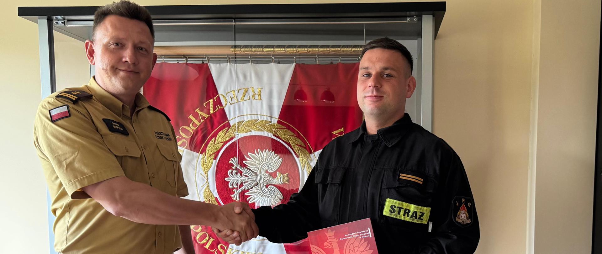 Na pierwszym planie Komendant Powiatowy Państwowej Straży Pożarnej w Rypinie bryg. Piotr Wypych oraz sekc. Dawid Dąbrowski podczas wręczania aktu mianowania na stanowisko ratownika. Strażacy podają sobie dłonie. W tle widoczny jest sztandar Komendy Powiatowej Państwowej Straży Pożarnej w Rypinie.a
