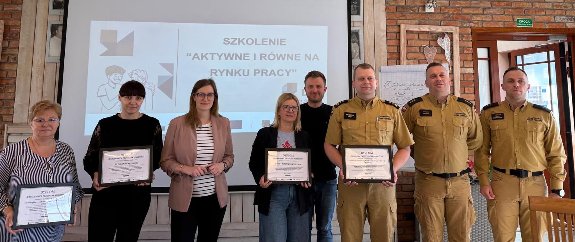 Uczestnicy szkolenia w tym funkcjonariusze PSP w żółtym umundurowaniu służbowym pozują do zdjęcia na tle telebimu.