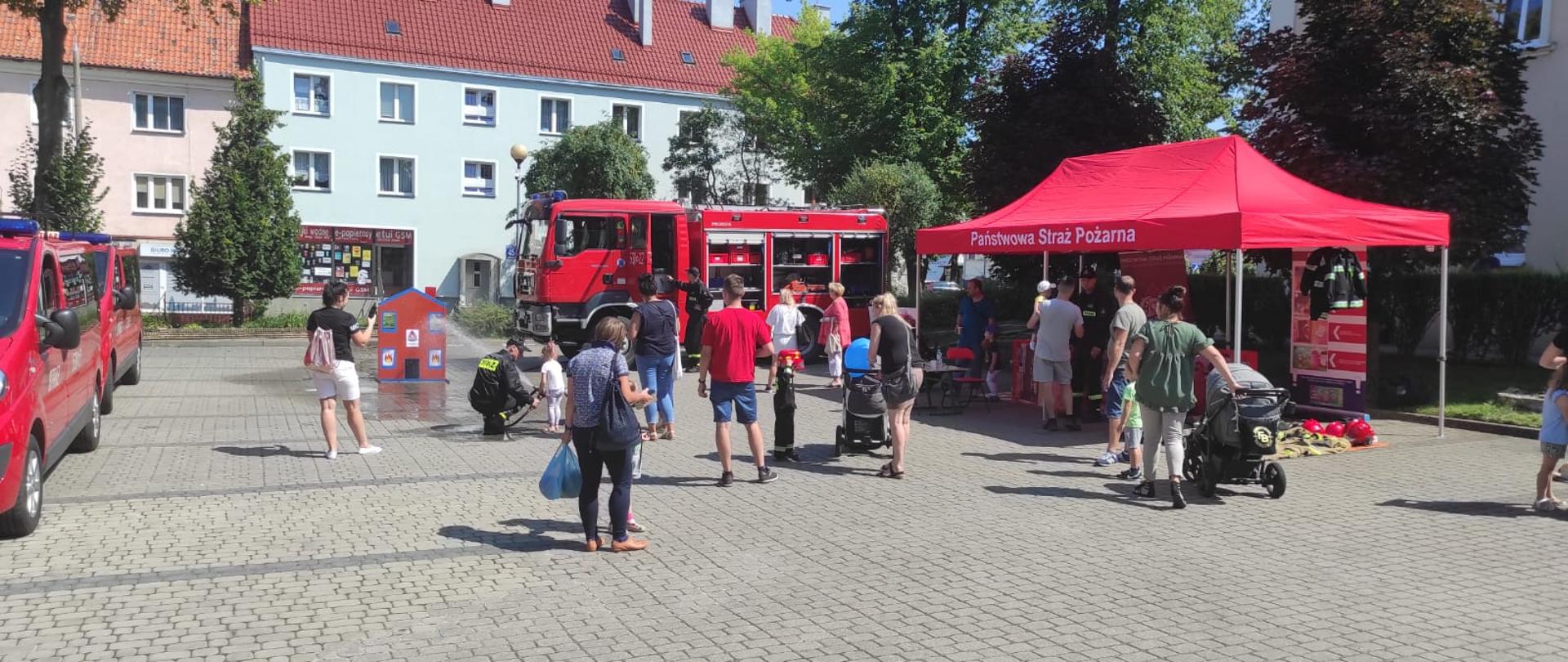 Grupa uczestników festynu przygląda się stanowisku Państwowej Straży Pożarnej. Pod czerwonym namiotem znajdują się banery informacyjne oraz stroje strażackie. W tle widać ciężarówkę strażacką, z której dzieci mogą podawać wodę do makiety palącego się domu.