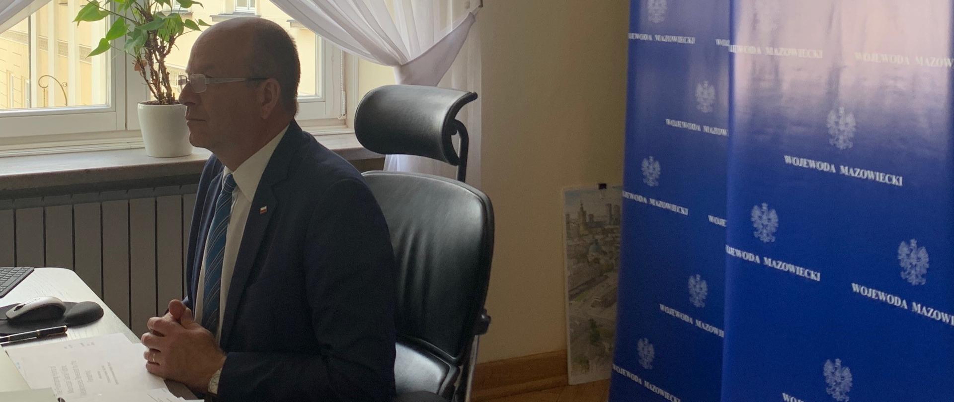 Na zdjęciu znajduje się Wojewoda Mazowiecki, który bierze udział w międzynarodowym kongresie. Z prawej strony znajdują się granatowe rool upy z logo Wojewody Mazowieckiego, a w tle okno