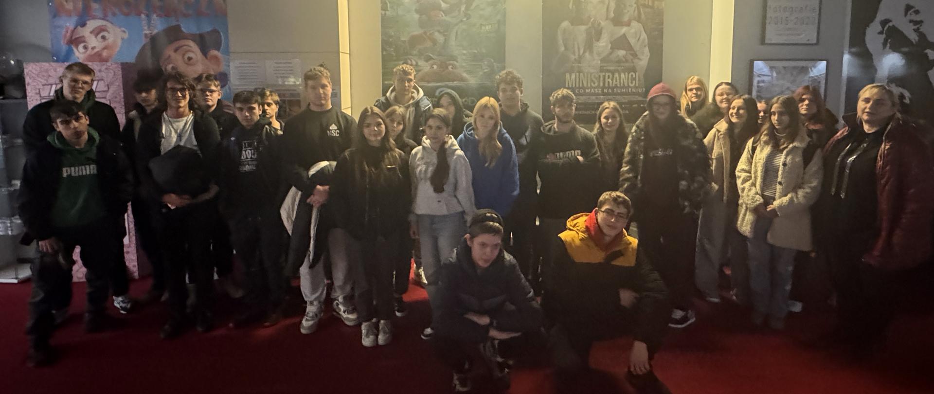 Grupa młodzieży stoi na czerwonym dywanie w holu kina przed plakatami filmowymi.