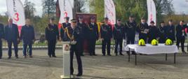 Pomorski komendant wojewódzki Państwowej Straży Pożarnej przemawia przez mikrofon stojąc przed mównicą za nim stoją zaproszeni goście oraz stoi stolik pokryty białym obrusem na którym położone są żółte hełmy.