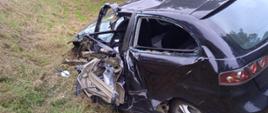 Na zdjęciu widoczny rozbity samochód osobowy marki Seat koloru ciemnego. Zdjęcie wykonane od strony lewego tyłu pojazdu. Na drugim planie widoczny na jezdni żółty ambulans i dwóch strażaków.