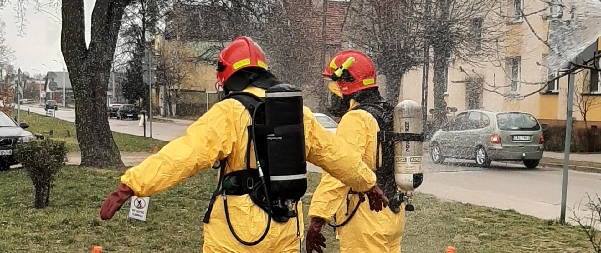 Zdjęcie przedstawia dwóch strażaków ubranych w żółte kombinezony. Na głowach mają czerwone hełmy, a na plecach czarne aparaty ochrony układu oddechowego. Osoby stoją między 3 pomarańczowymi pachołkami na trawie. W tle widać budynki mieszkalne oraz drzewa.