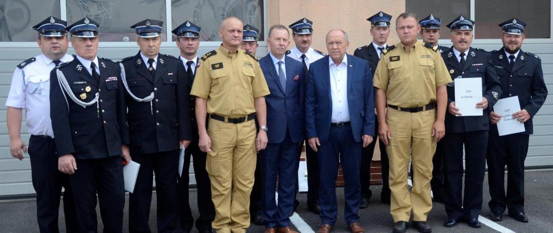 Przed budynkiem Jednostki Ratowniczo – Gaśniczej w Kolbuszowej na tle bram garażowych stoi grupa kilkunastu osób: dwóch strażaków PSP w mundurach w kolorze piaskowym, strażacy OSP w mundurach galowych oraz osoby cywilne w garniturach.