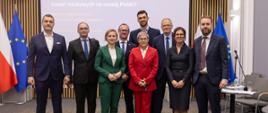Pod wbudowanym w ścianę z cienkich pionowych listewek wielkim ekranem stoi dziewięć osób, drugi od lewej minister Kulasek trzyma szarą teczkę, za nimi pod ścianą po obu stronach flagi Polski i UE.