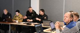 Strażak dyskutuje z drugim strażakiem obok siedzą przedstawiciele instytucji przed laptopami podczas ćwiczenia w formie sztabowej.